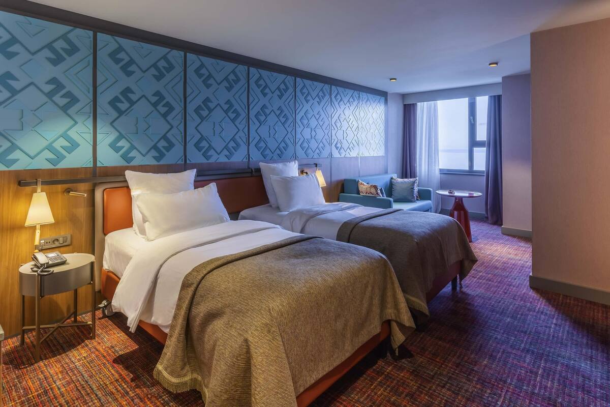 Ein Hotelzimmer mit zwei Betten, einem kleinen Tisch, einer Lampe und einem Fenster mit Vorhängen.