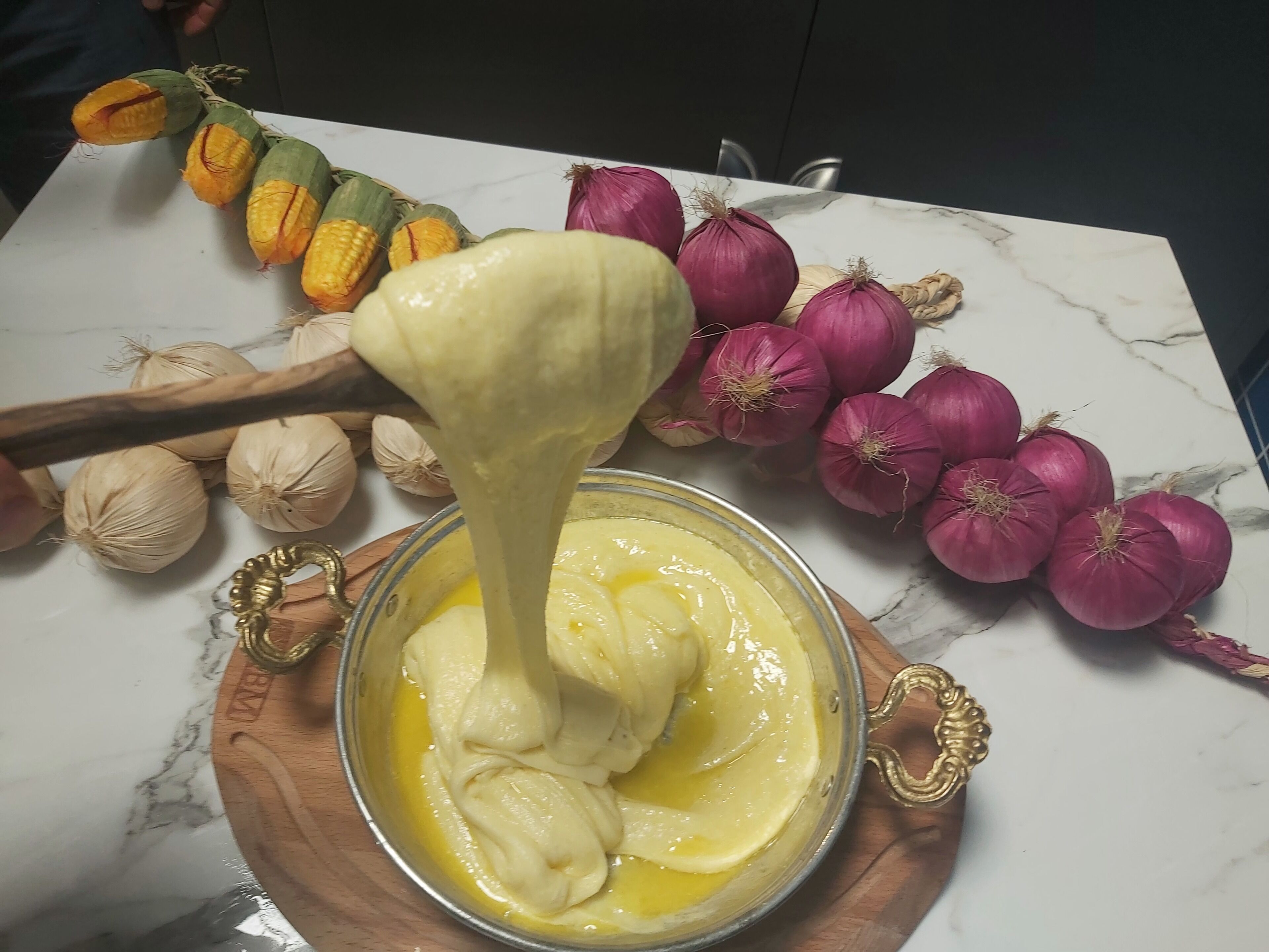 A bowl of yellow sauce with a ladle, surrounded by onions and garlic on a marble countertop.