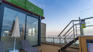 Una terraza con pared de vidrio, una silla de lounge, una sombrilla y una escalera que conduce a una cubierta superior.