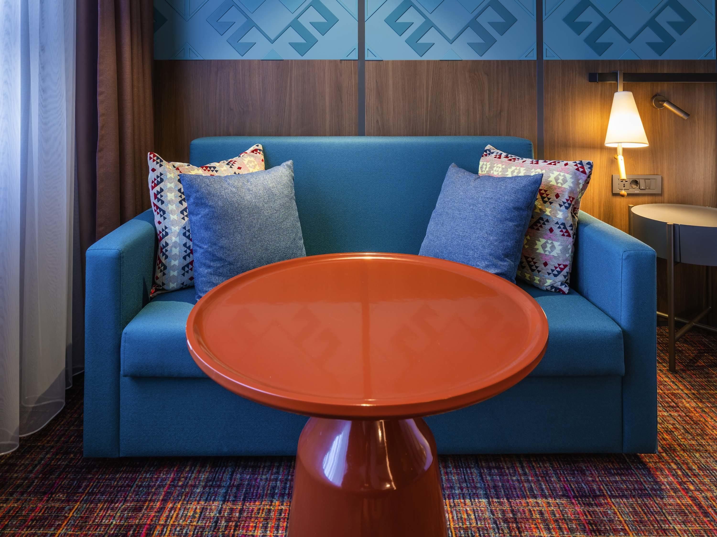 A blue sofa with patterned pillows, a round orange table, and a lamp on a wooden wall.