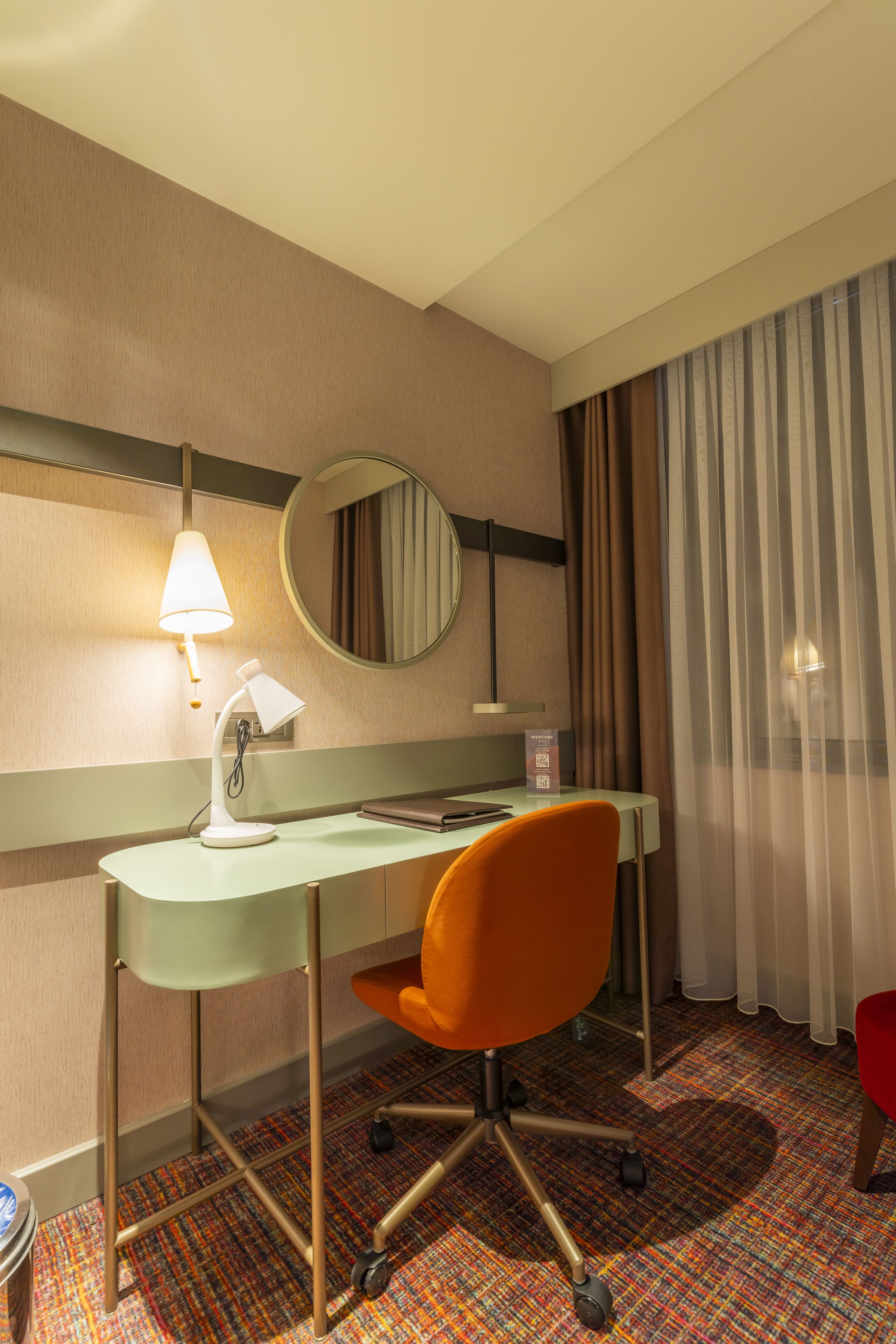 Habitación de hotel con un escritorio, una silla de oficina color naranja, un espejo redondo, una lámpara y una ventana con cortinas.