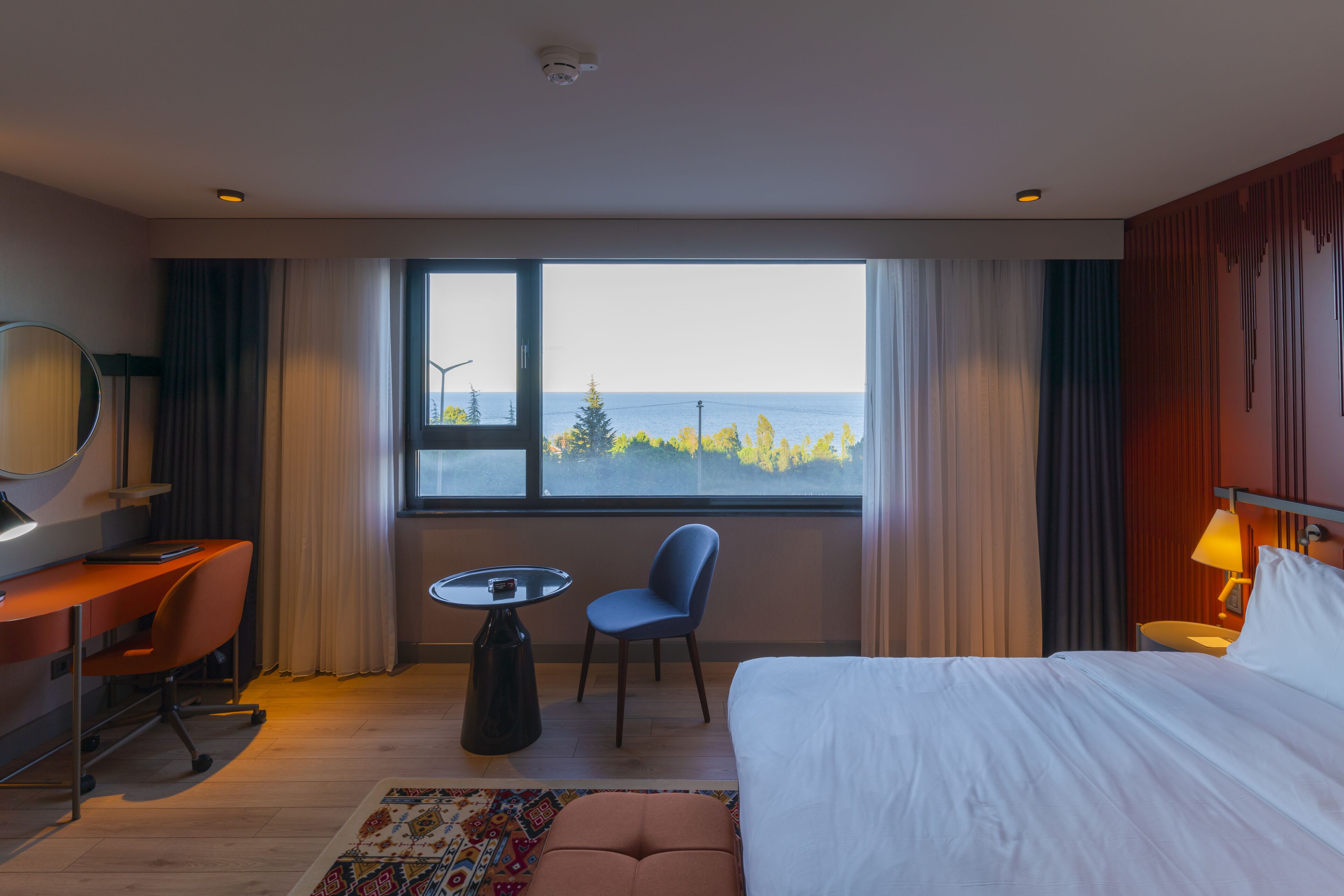 A hotel room with a bed, desk, chair, and a view of the sea.