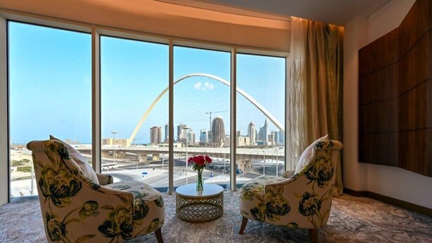 Una habitación con dos sillones estampados, una mesa redonda con un jarrón de flores rojas y amplias ventanas que ofrecen vistas a un paisaje urbano con un puente.