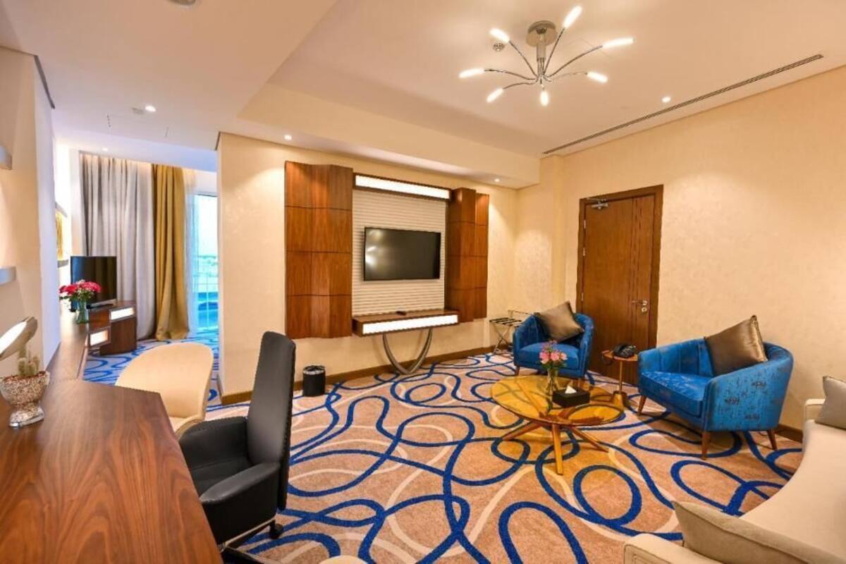 A hotel room with a wooden desk, two sofas, a TV, and a patterned carpet.