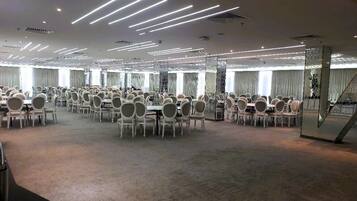 Un amplio salón de conferencias con filas de sillas y mesas blancas, iluminación moderna y ventanas grandes con cortinas.