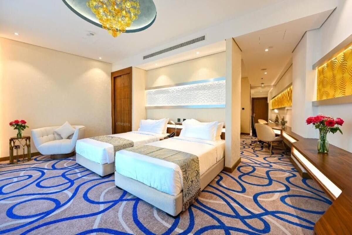 A hotel room with two beds, a desk, a chair, a small table with flowers, and a decorative wall panel.
