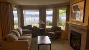 A living room with a sofa, armchair, and fireplace, large windows with a view of a lake. - Oceanfront Nanoose Bay stunning views condo sleeps 6 (Nanoose Bay)