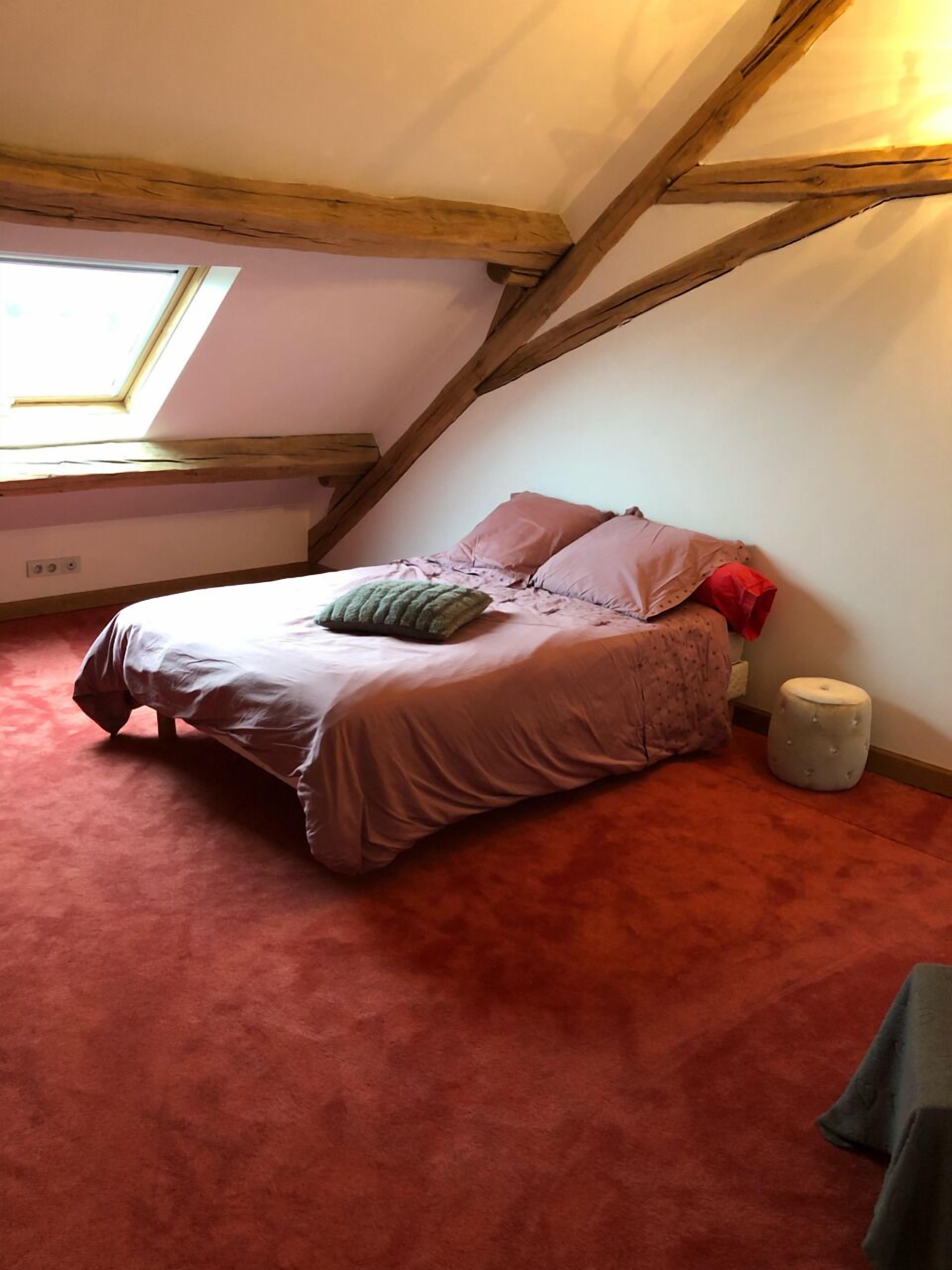 A bedroom with a sloped ceiling, a bed with a pink bedspread and pillows, a small stool, and a skylight.