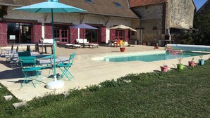 Ein Poolbereich mit Liegestühlen und Sonnenschirmen, ein Steinhaus mit Ziegeldach und ein grasbewachsenes Vordergrund.