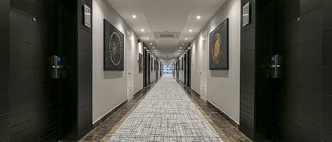 A long, narrow hotel corridor with dark walls, a patterned carpet, and framed artwork on the walls.