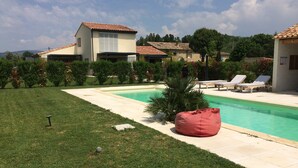 A swimming pool with a red bean bag chair, a grassy area, and a house with a tiled roof. - 4* apartment facing the Luberon (Oppède)