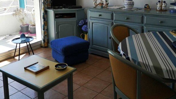 Una sala de estar con televisor, una pequeña decoración de velero, un cuadro de peces y un comedor con un sofá azul.