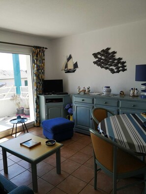 A living room with a TV, a small sailboat decoration, a wall art of fish, a dining table, and a blue sofa.