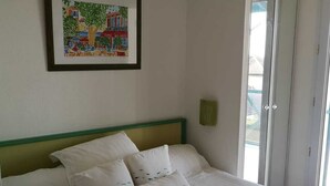 Una cama bien tendida con sábanas blancas y almohadas decorativas, un cuadro en la pared y un ventanal corredero a la derecha.