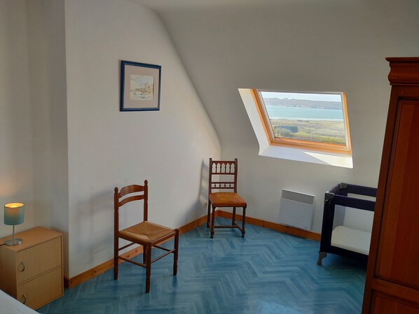 A room with a sloped ceiling, a wooden chair, a small cabinet, and a window with a view of a landscape.