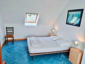 A bedroom with a bed, a chair, a nightstand, a small cabinet, a window, and a framed picture on the wall.