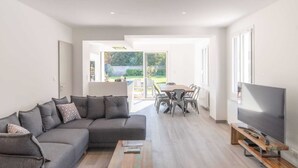 Un salon moderne comprenant un canapé d’angle, une table basse en bois, un coin repas avec une table et des chaises, et un téléviseur posé sur un meuble en bois.