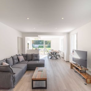 A modern living room with a sectional sofa, a wooden coffee table, a dining area with a table and chairs, and a television on a wooden stand.