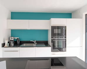 A modern kitchen with white cabinets, a teal accent wall, a stainless steel sink, a coffee maker, and a built-in oven.