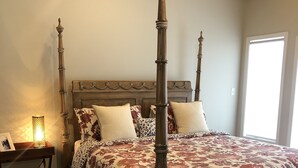 Een hemelbed met een bloemenlaken, een houten nachtkastje met een lamp, en een zacht vloerkleed op een houten vloer.