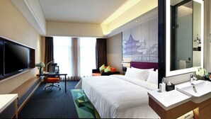 Habitación de hotel con cama, escritorio con silla, televisor, baño con lavamanos y espejo, y un ventanal con cortinas.
