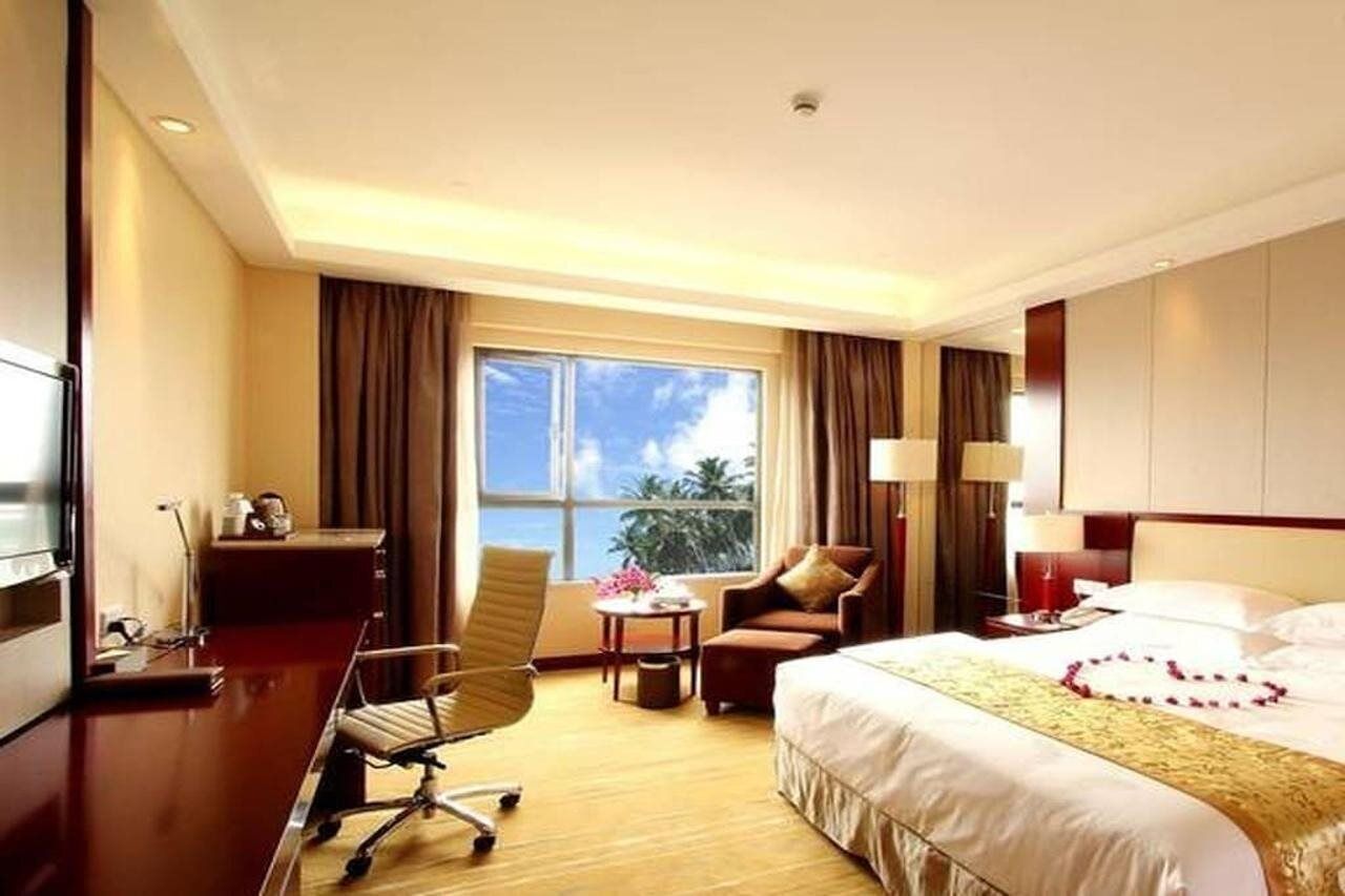 A hotel room with a bed, a desk, a chair, a small table with flowers, and a view of the outdoors.