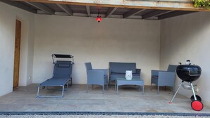 En uteplats med ett takförsett område, en solstol, en soffa, ett litet bord med en dekorativ detalj och en grill.