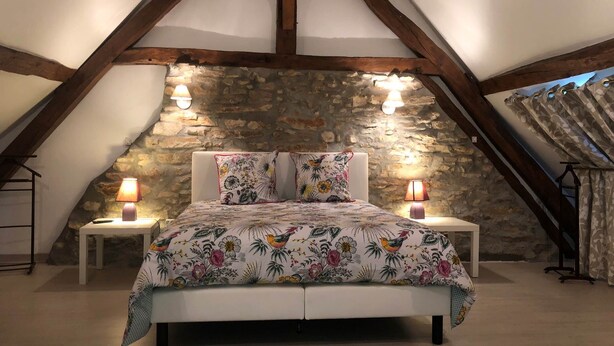 Een slaapkamer met een schuin dak, zichtbare houten balken, een stenen muur en een bed met bloemenbeddengoed.