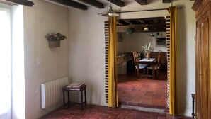 Een rustieke kamer met zichtbare houten balken, een tegelvloer van baksteen, een eettafel met stoelen, en een keuken die zichtbaar is door een open deur.
