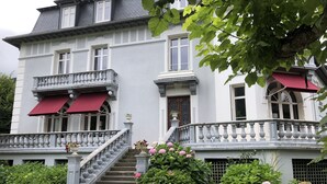 A two-story building with a balcony, a staircase, and a garden with pink flowers.