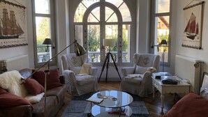 A cozy living room with two armchairs, a sofa, a coffee table, and a lamp. Large windows with a view of greenery are visible.