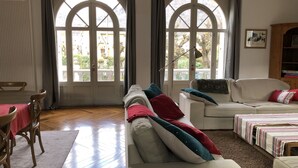 A living room with a wooden floor, two white sofas, a dining table with chairs, and large arched windows with curtains.