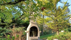 A stone-built outdoor fireplace with logs stored underneath, surrounded by gravel and greenery.