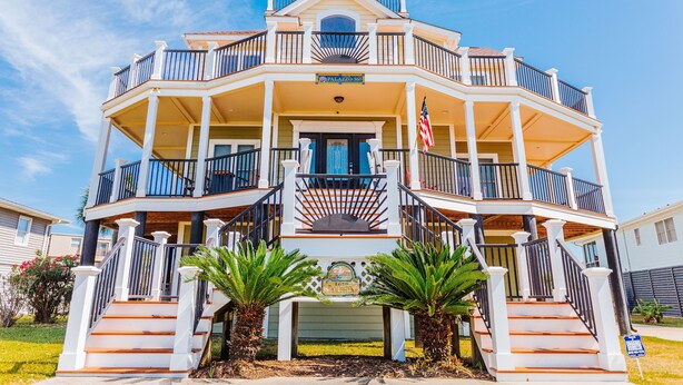A multi-story building with a balcony on each floor, a central entrance with a decorative sign, and an American flag. - Palazzo 360 5bdr/5 bath with a heated pool sleeps 18 (Murrells Inlet)