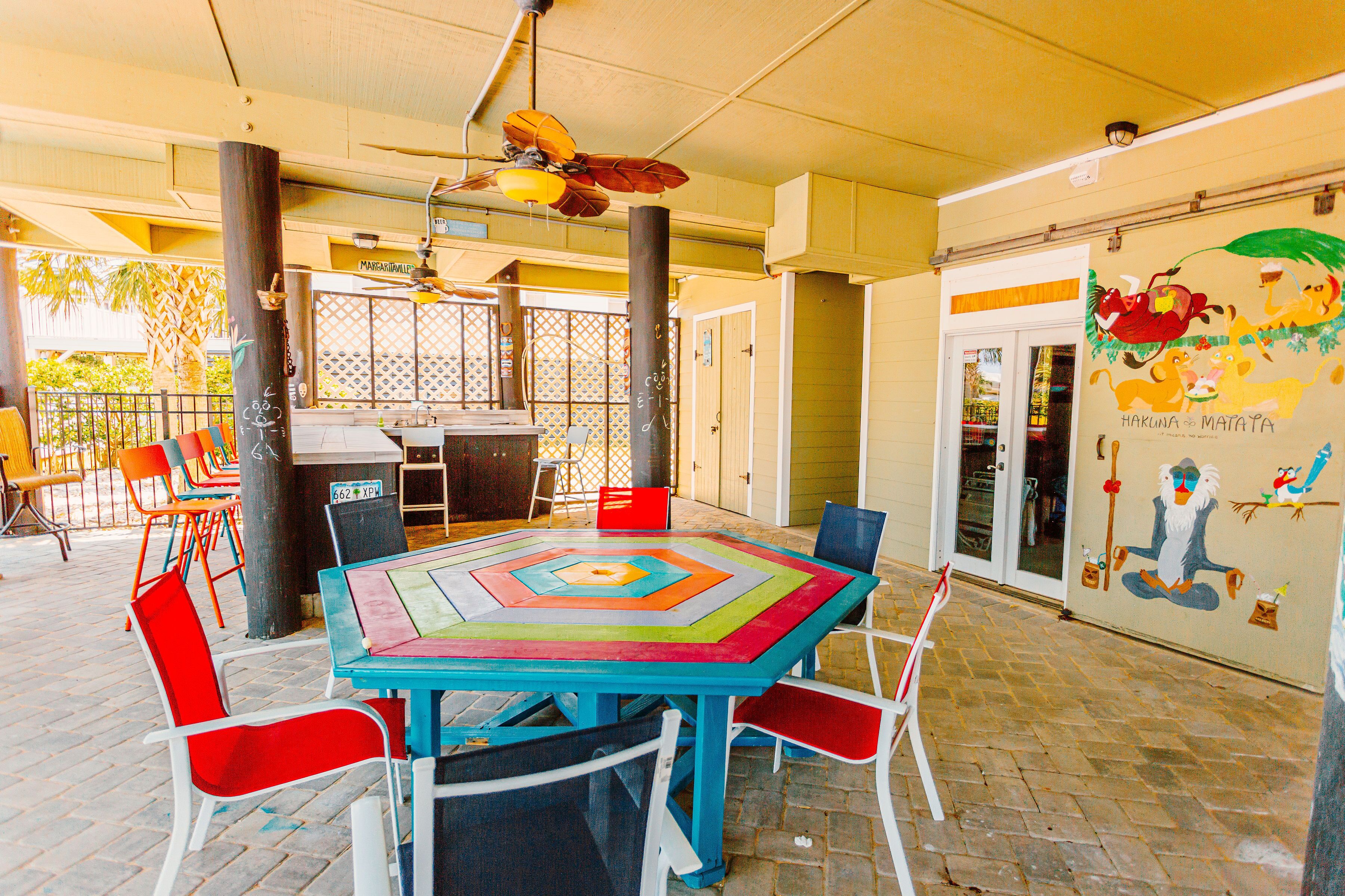 An outdoor dining area with a colorful table and chairs, a ceiling fan, and a mural on the wall.