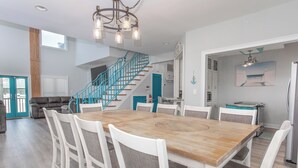 A spacious dining area with a wooden table and white chairs, a staircase with teal railings, and a kitchen area with a refrigerator and microwave. - Palazzo 360 5bdr/5 bath with a heated pool sleeps 18 (Murrells Inlet)