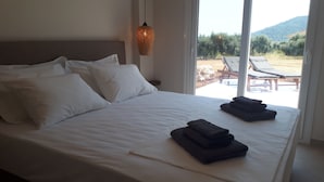 Een net opgemaakt bed met witte lakens en kussens, een geweven lamp boven, en uitzicht op zitplaatsen buiten en groen door grote ramen.