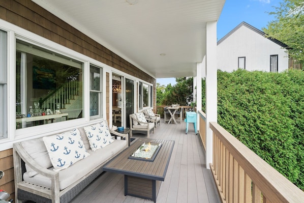 A covered outdoor seating area with a wooden bench and a table, surrounded by large windows and a hedge.