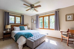 Une chambre à coucher comprenant un lit, des tables de chevet, une chaise, un ventilateur de plafond et de grandes fenêtres.