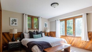 Un dormitorio con cama, mesitas de noche, un banco y dos ventanas con cortinas.