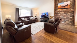 A modern living room with a sectional sofa, leather armchairs, a flat-screen TV, and a brick fireplace.