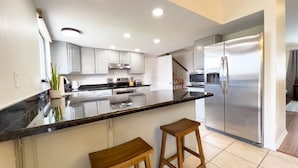 A modern kitchen with white cabinets, a black countertop, stainless steel appliances, and two wooden stools.