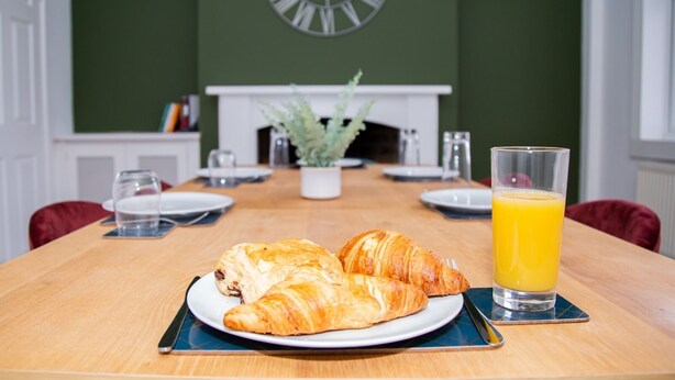 Une table mise pour le petit-déjeuner avec des croissants et du jus d’orange, une horloge au mur et une cheminée en arrière-plan.