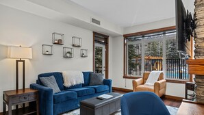 Un salon avec un canapé bleu, une table basse, une lampe et un fauteuil. Les murs sont ornés de tableaux encadrés, et une fenêtre offre une vue sur des arbres et une piscine.
