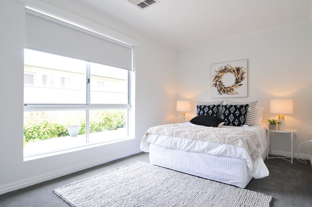 A bedroom with a large window, a bed with white bedding, two bedside lamps, and a wall-mounted artwork.