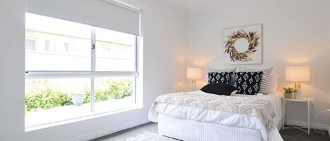 A bedroom with a large window, a bed with white bedding, two bedside lamps, and a wall-mounted artwork.