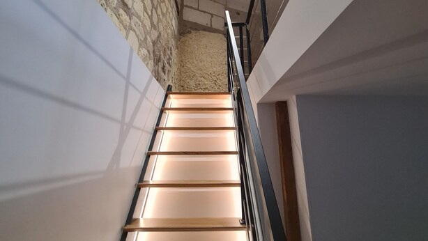Una escalera con escalones de madera y barandilla metálica, que conduce a una habitación con pared de piedra y viga de techo de madera.