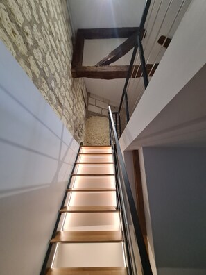 A staircase with wooden steps and a metal railing, leading to a room with a stone wall and a wooden ceiling beam.
