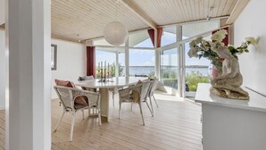 Zona pranzo con tavolo rotondo, sedie in vimini e vista sul mare attraverso ampie finestre.