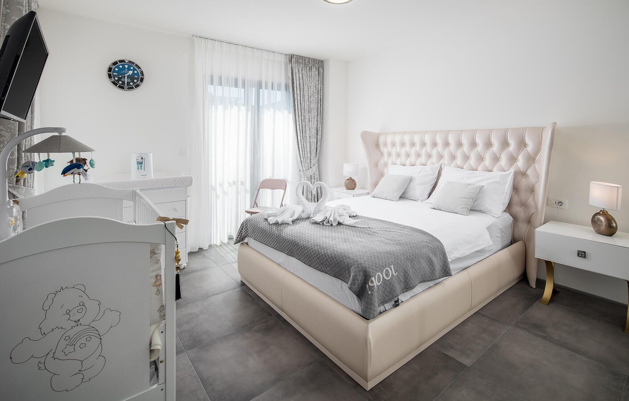 A modern bedroom with a large bed, a bedside table, a dresser, a TV, and a clock on the wall.